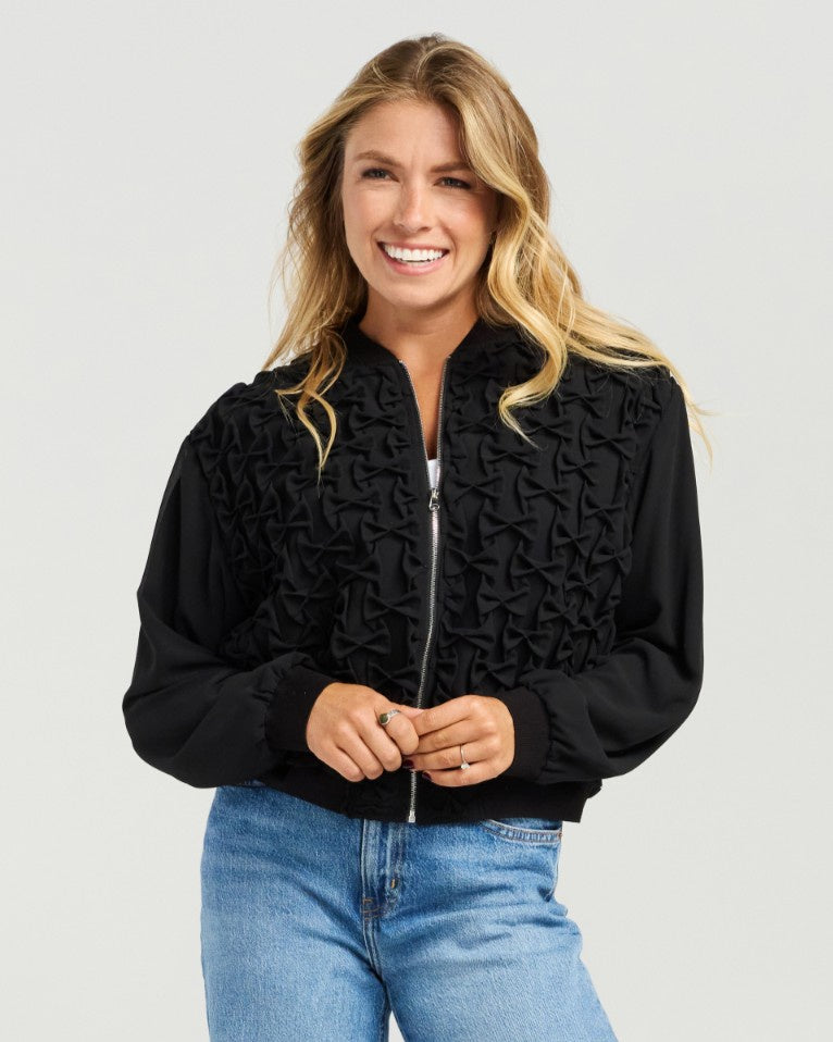 A smiling blonde woman wearing a black textured zip-up bomber jacket with long sleeves and blue jeans, standing against a light gray studio background.