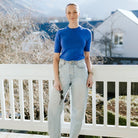 A woman stands on a white porch wearing a bright royal blue short-sleeve crewneck t-shirt and light-wash high-waisted wide-leg jeans, holding sunglasses in one hand with a scenic outdoor background.