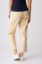 A person shown from the back wearing light beige cropped trousers with a small embroidered logo on the back pocket, paired with a dark navy top and white sneakers, against a light gray studio background.