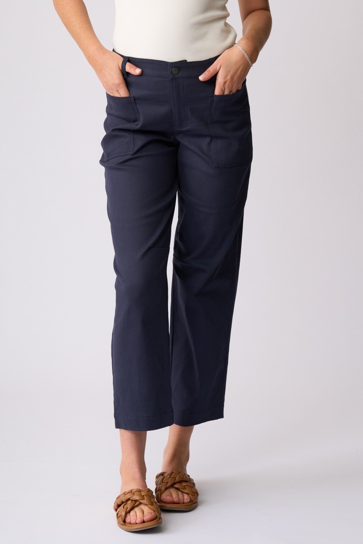 A person wearing navy cropped pants with a cream top and brown sandals, standing against a light gray studio background.