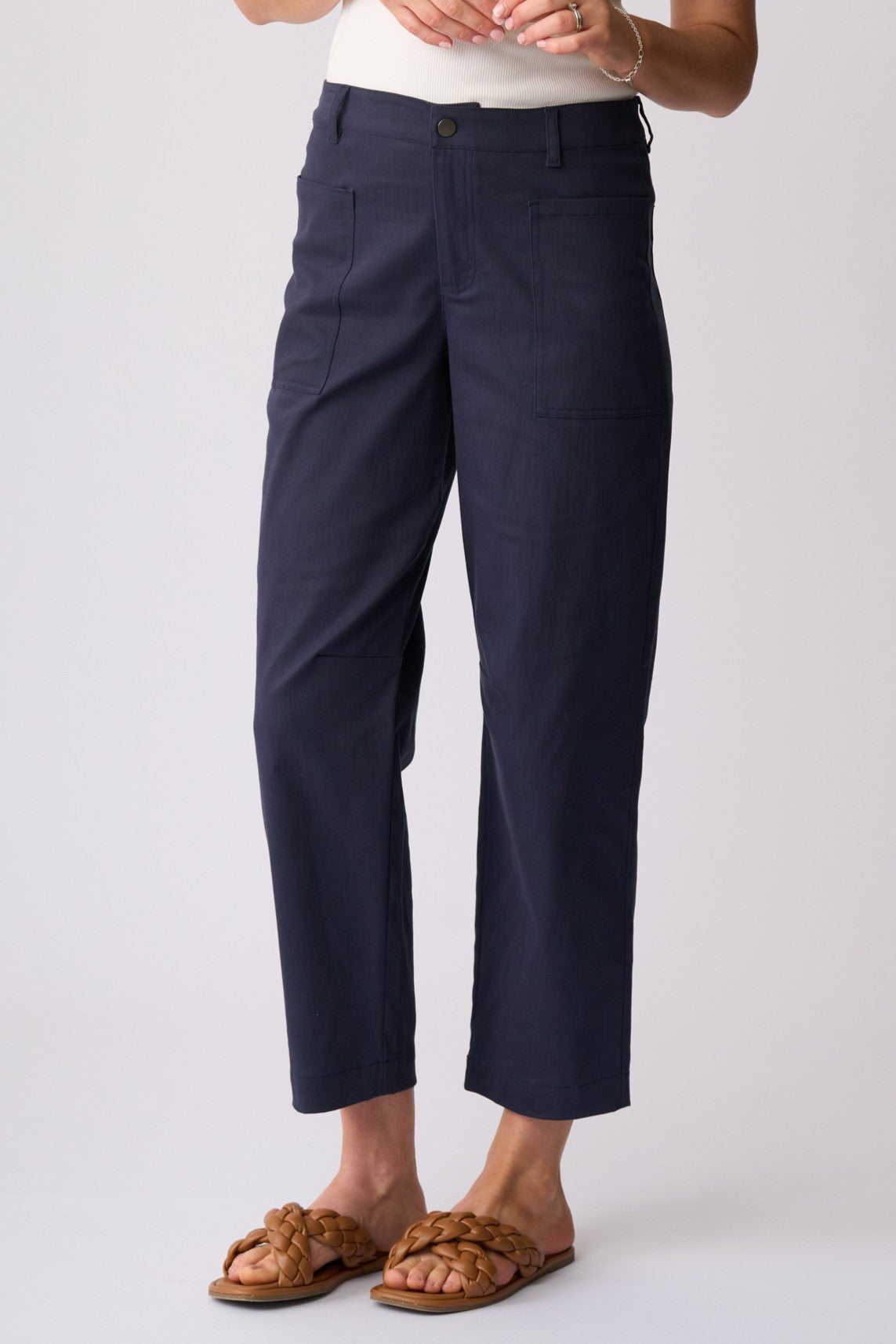 A person wearing navy cropped pants with a white top and brown woven slide sandals, shown from the waist down against a light gray studio background.
