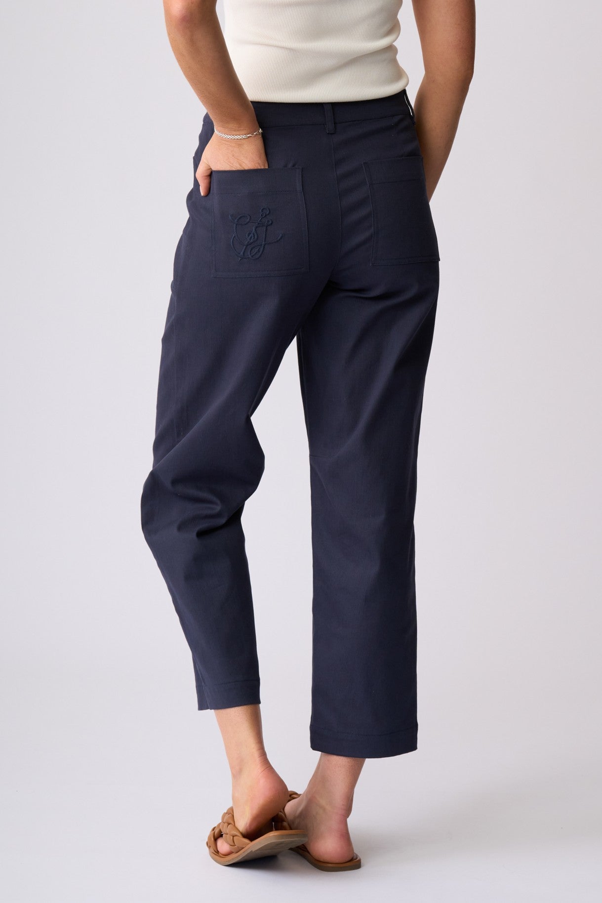 A person shown from the back wearing navy cropped pants with a back pocket and embroidered logo detail, paired with a cream top and tan sandals, against a light gray studio background.