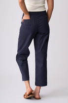 A person shown from the back wearing navy cropped pants with a back pocket and embroidered logo detail, paired with a cream top and tan sandals, against a light gray studio background.