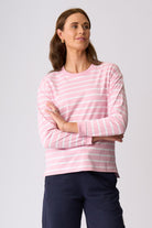 A woman modeling a pink and white striped long-sleeve crewneck top with dark navy pants, standing against a light gray studio background.