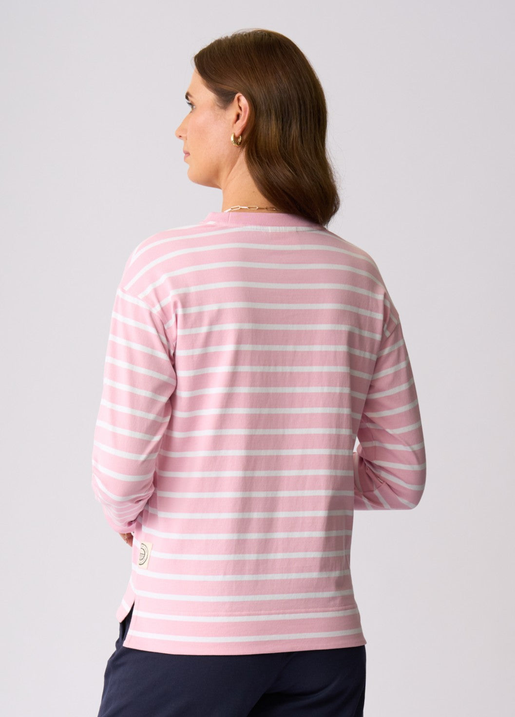 A woman shown from the back wearing a pink and white horizontally striped long-sleeve top, paired with dark navy pants, against a light gray studio background.