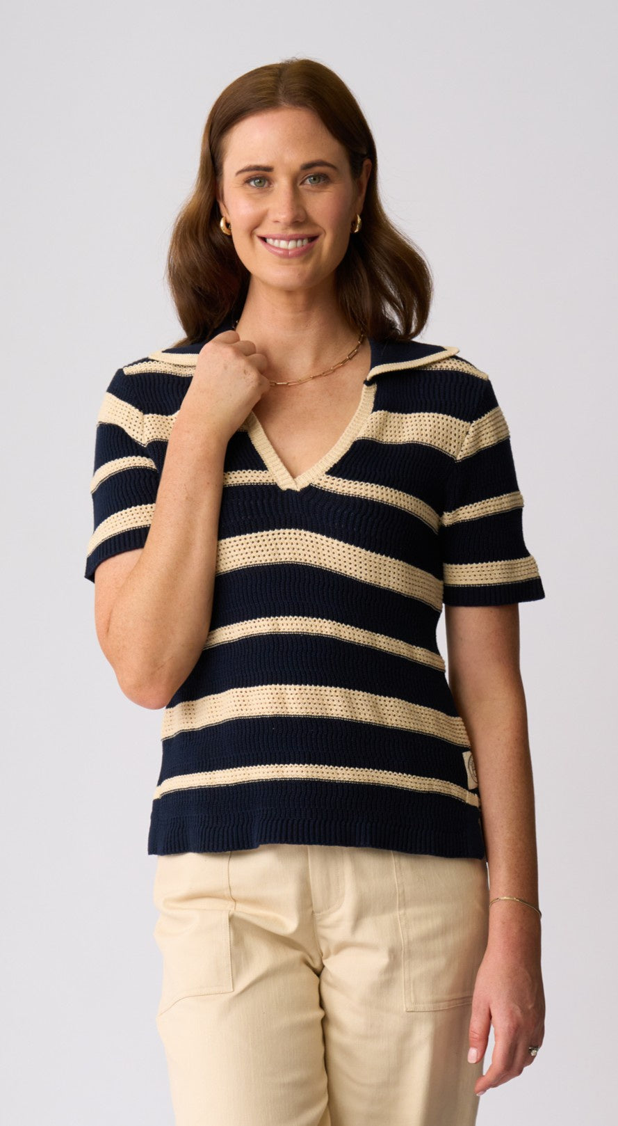 A woman wearing a short-sleeve knit V-neck top with navy and beige horizontal stripes, paired with light beige pants, against a light gray studio background.