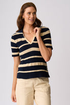 A woman wearing a navy and cream striped short-sleeve knit polo shirt with beige pants, standing against a light gray studio background.