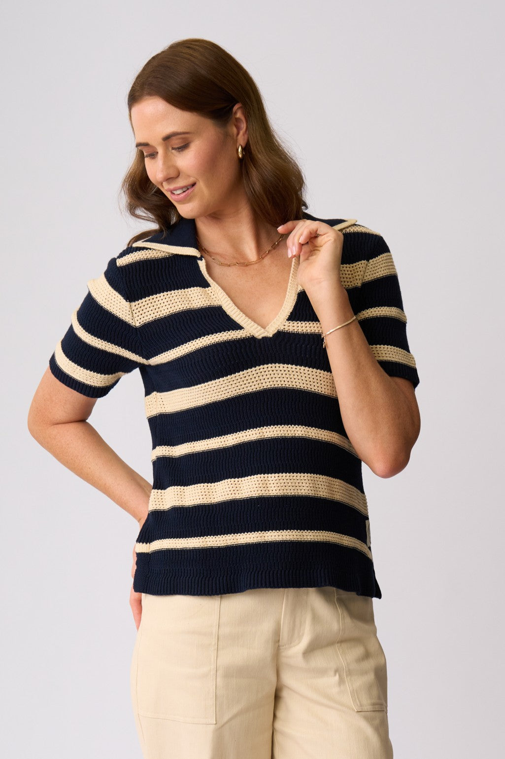 A woman wearing a short-sleeve knit polo-style top with horizontal navy and cream stripes, paired with light beige pants, photographed against a plain light gray studio background.