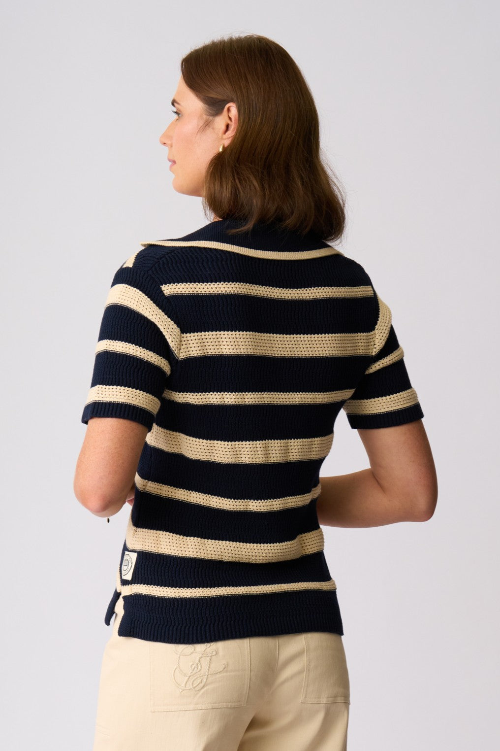 A woman shown from the back wearing a short-sleeve knit polo shirt with navy and beige horizontal stripes, paired with beige trousers, against a light gray studio background.