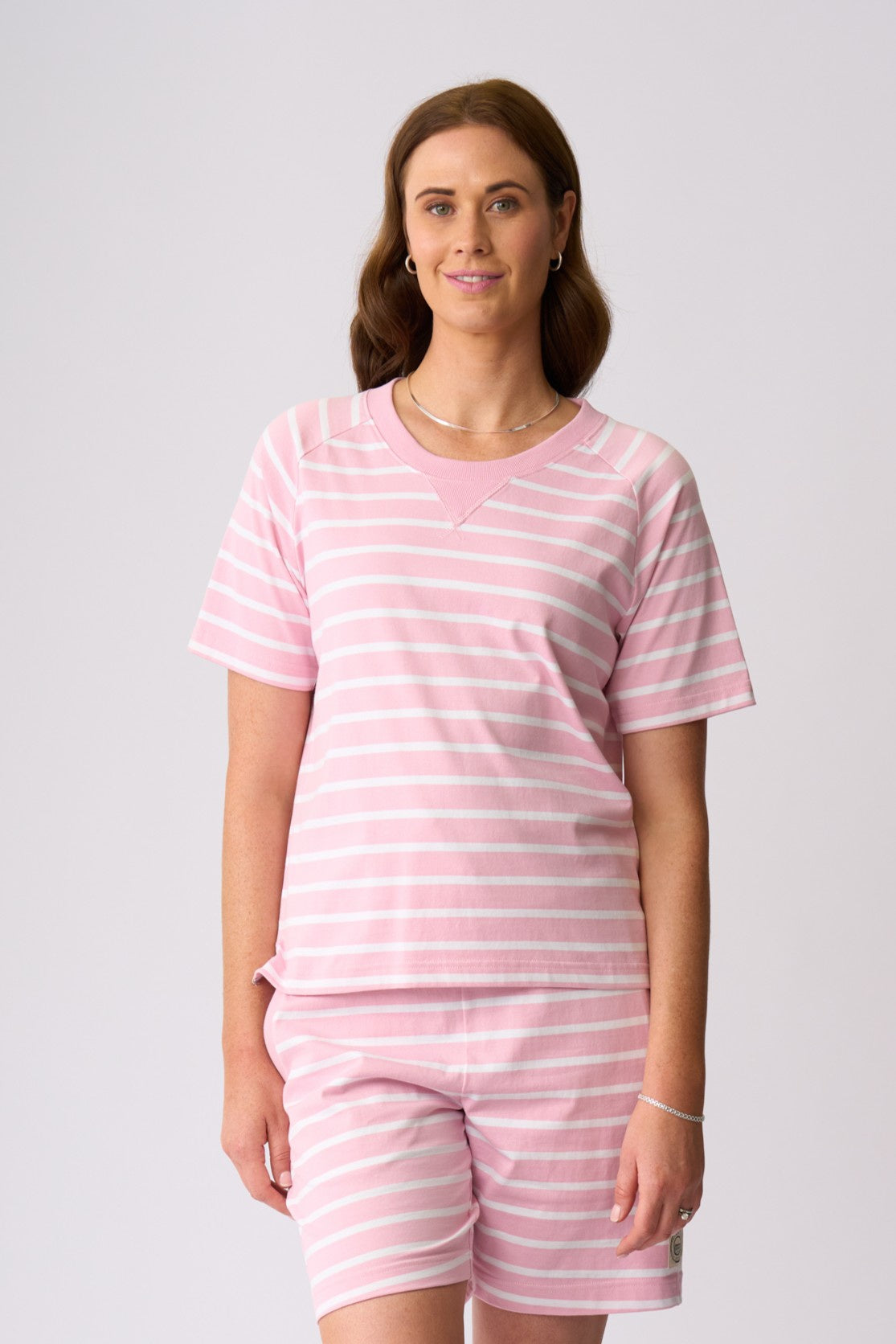 A woman modeling a matching two-piece loungewear set consisting of a short-sleeve V-neck top and matching shorts, both in a light pink and white horizontal stripe pattern, against a solid light gray background.