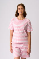 A woman modeling a matching two-piece loungewear set consisting of a short-sleeve V-neck top and matching shorts, both in a light pink and white horizontal stripe pattern, against a solid light gray background.