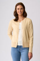 A woman modeling a light beige cardigan with scalloped edges and decorative trim over a white top and blue jeans, photographed against a plain light gray studio background.