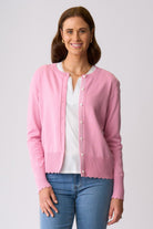 A woman modeling a light pink knit cardigan with scalloped edges and a decorative chevron pattern on the placket, worn over a white top and blue jeans against a light gray studio background.