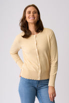 A woman modeling a light beige cardigan sweater with scalloped edges and white buttons, paired with blue jeans, against a solid light gray background.