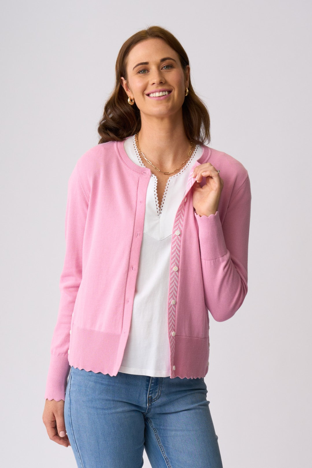 A woman modeling a light pink knit cardigan over a white top and blue jeans, photographed against a plain light gray studio background.