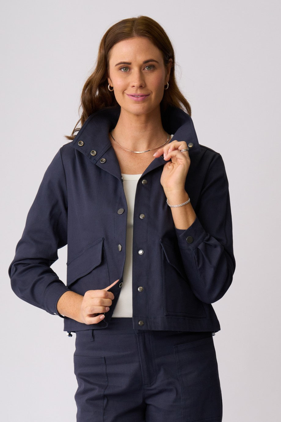 A woman modeling a navy blue cropped utility jacket with a high collar, snap buttons, and front flap pockets, paired with matching navy pants against a light gray studio background.