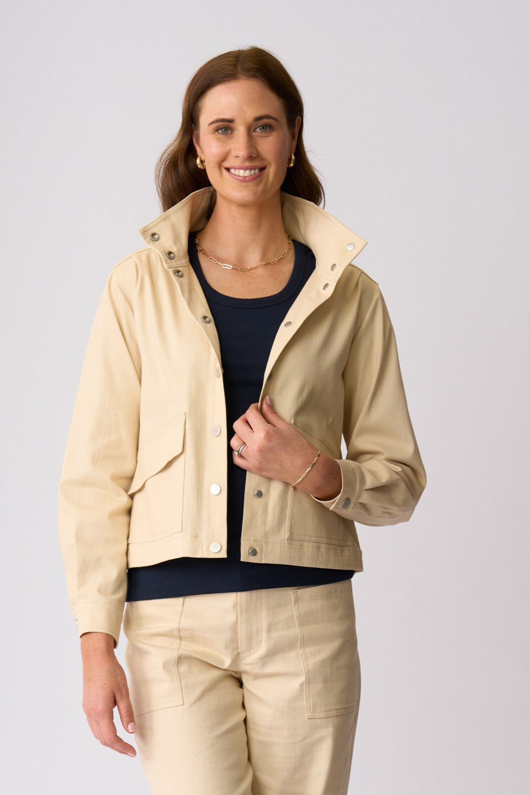A woman modeling a light beige utility jacket with a stand-up collar and snap-button front, worn over a dark navy top and paired with matching beige pants, against a light gray studio background.