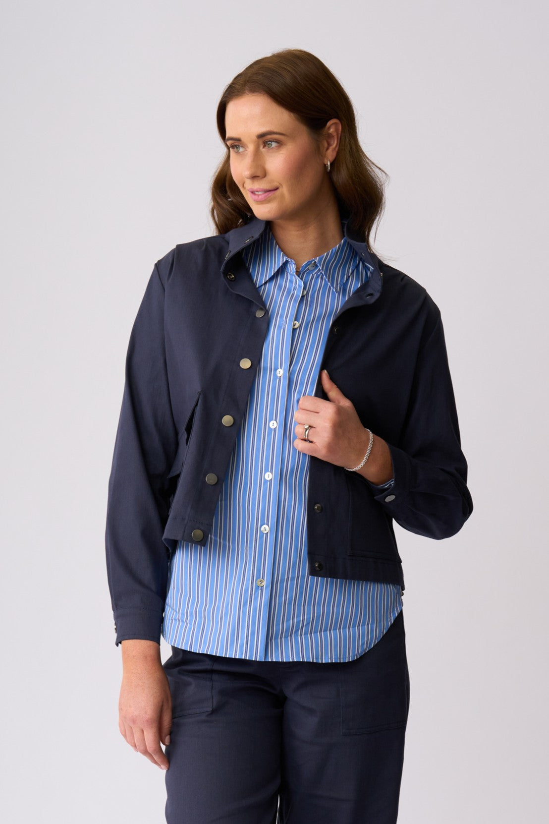 A woman modeling a navy blue utility jacket over a blue and white striped button-down shirt, paired with navy trousers, against a light gray studio background.