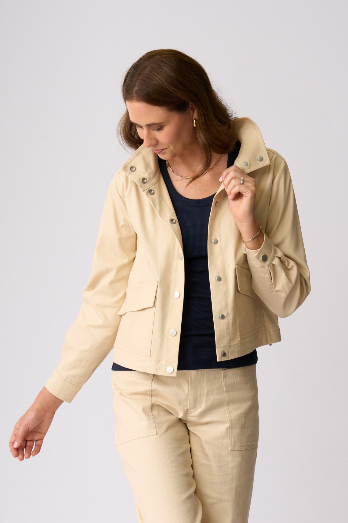 A woman wearing a light beige cropped utility jacket with silver snap buttons, matching beige trousers, and a dark navy top, photographed against a neutral light gray studio background.
