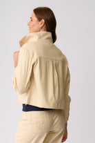 A woman shown from the back wearing a light beige cropped jacket and matching beige trousers against a plain light gray studio background.