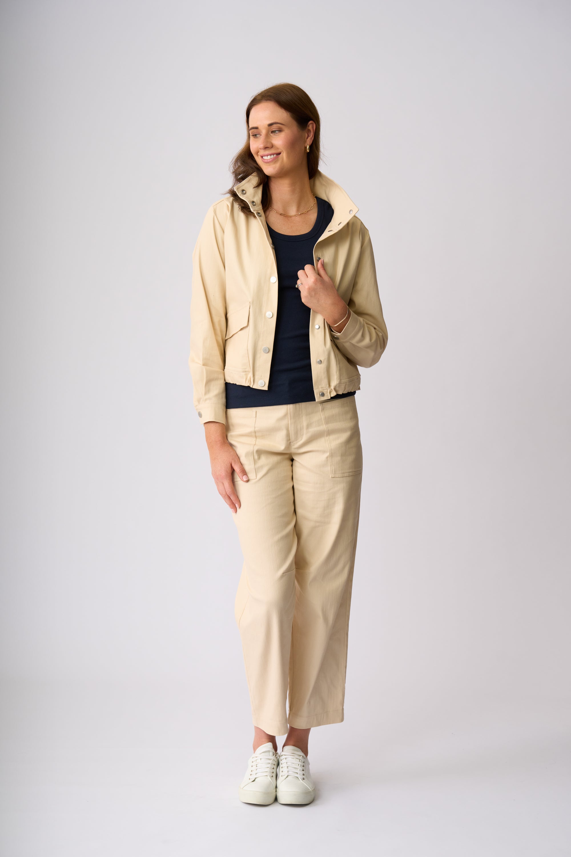A woman modeling a light beige cropped utility jacket, matching wide-leg trousers, a dark navy top, and white sneakers against a light gray studio background.