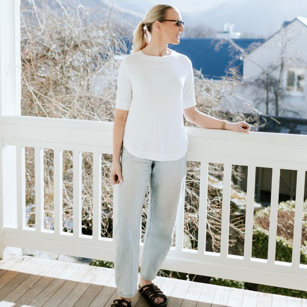 A woman wearing a white short-sleeve crewneck t-shirt and light-wash wide-leg jeans stands on a wooden deck leaning on a white railing, with a mountain and house visible in the background.