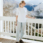 A woman wearing a white short-sleeve crewneck t-shirt and light-wash wide-leg jeans stands on a wooden deck leaning on a white railing, with a mountain and house visible in the background.