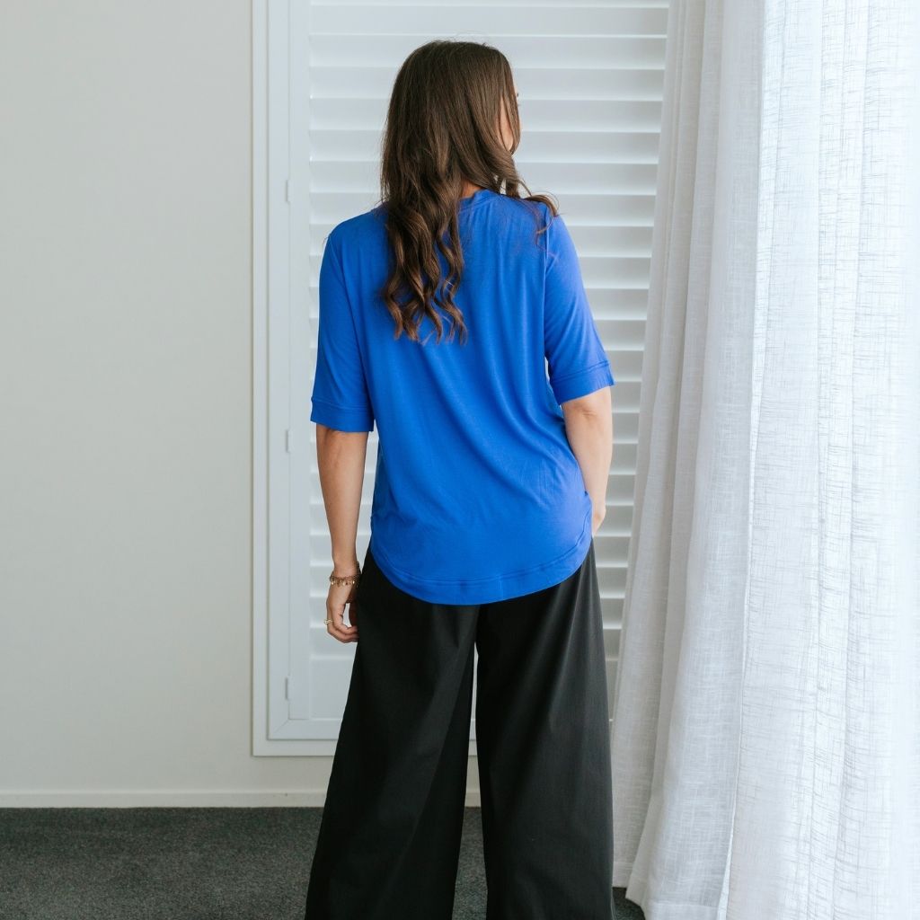 A woman viewed from behind wearing a bright blue short-sleeve top and black wide-leg pants, standing indoors near white window shutters and a sheer curtain.