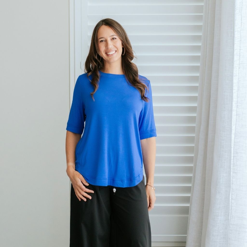 A woman wearing a bright blue short-sleeve top and black pants stands in front of white window blinds and a sheer curtain.