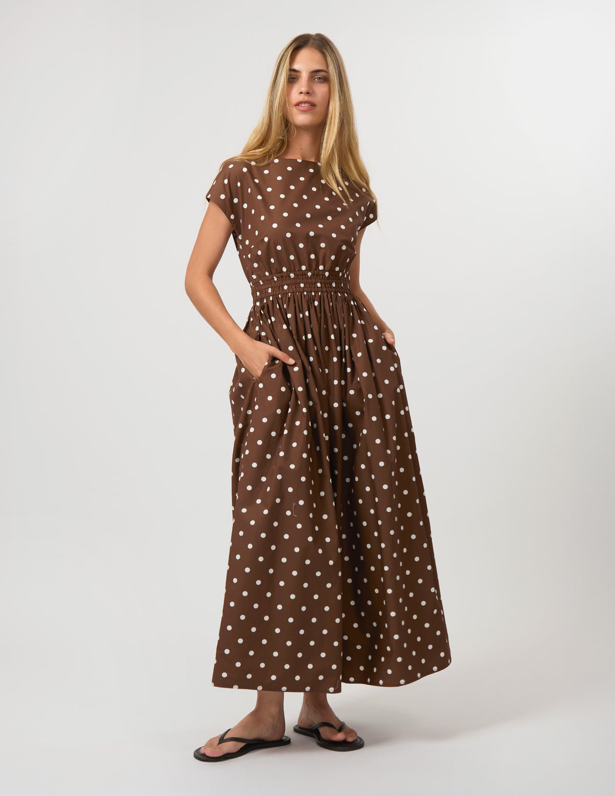 A woman modeling a brown short-sleeve maxi dress with a white polka dot pattern, standing against a plain light gray studio background.