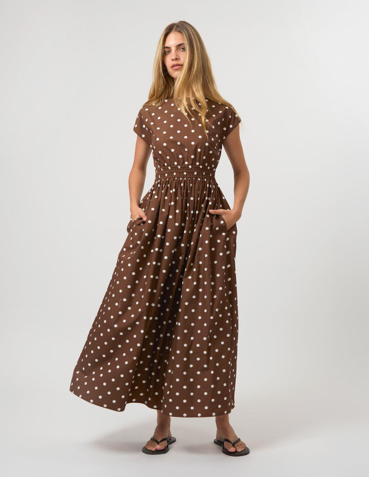 A woman modeling a brown short-sleeve maxi dress with white polka dots, standing against a light gray studio background.