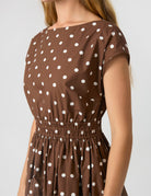 A close-up of a woman wearing a brown short-sleeve dress with a white polka dot pattern and an elasticized waist, shown against a light gray studio background.