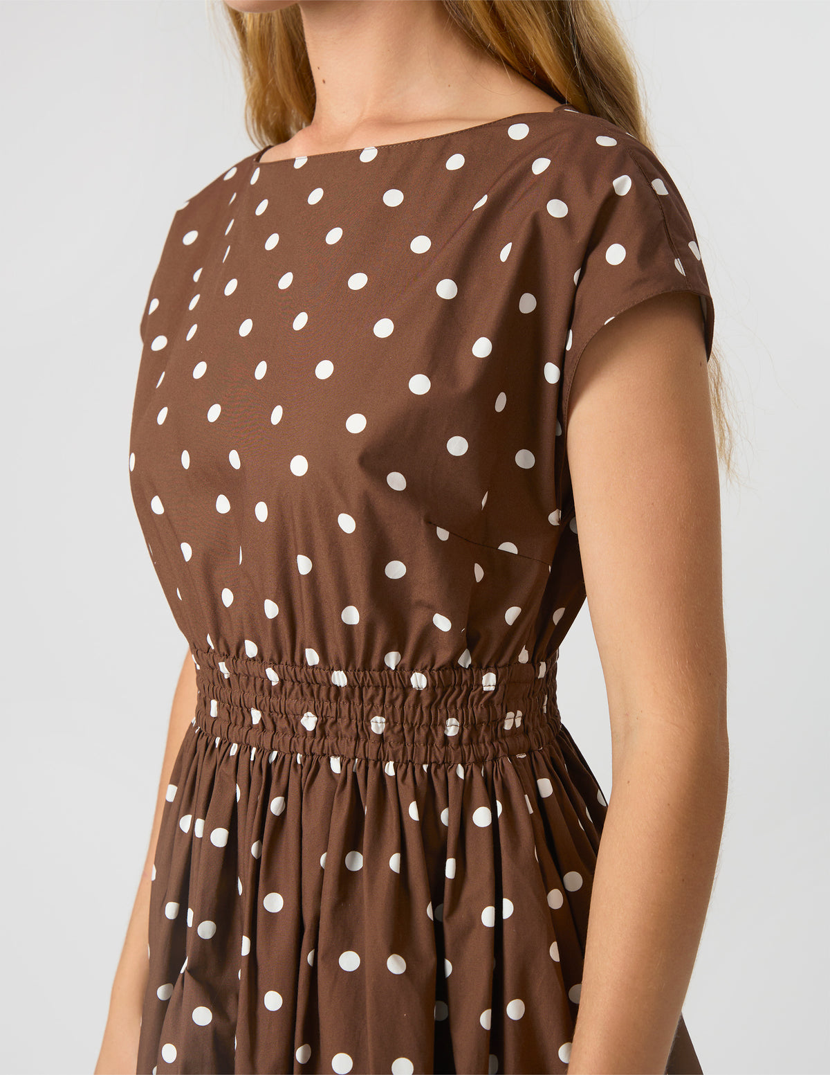 A close-up of a woman wearing a brown short-sleeve dress with a white polka dot pattern and an elasticized waist, shown against a light gray studio background.