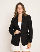 A woman modeling a black blazer over a white top and white shorts, standing against a plain light background.