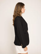 A woman shown in side profile wearing a black blazer with gold buttons and a white skirt, photographed against a plain off-white studio background.