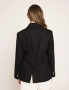 A woman viewed from the back wearing a black tailored blazer with gold-tone buttons on the cuffs and white pants, against a plain light background.