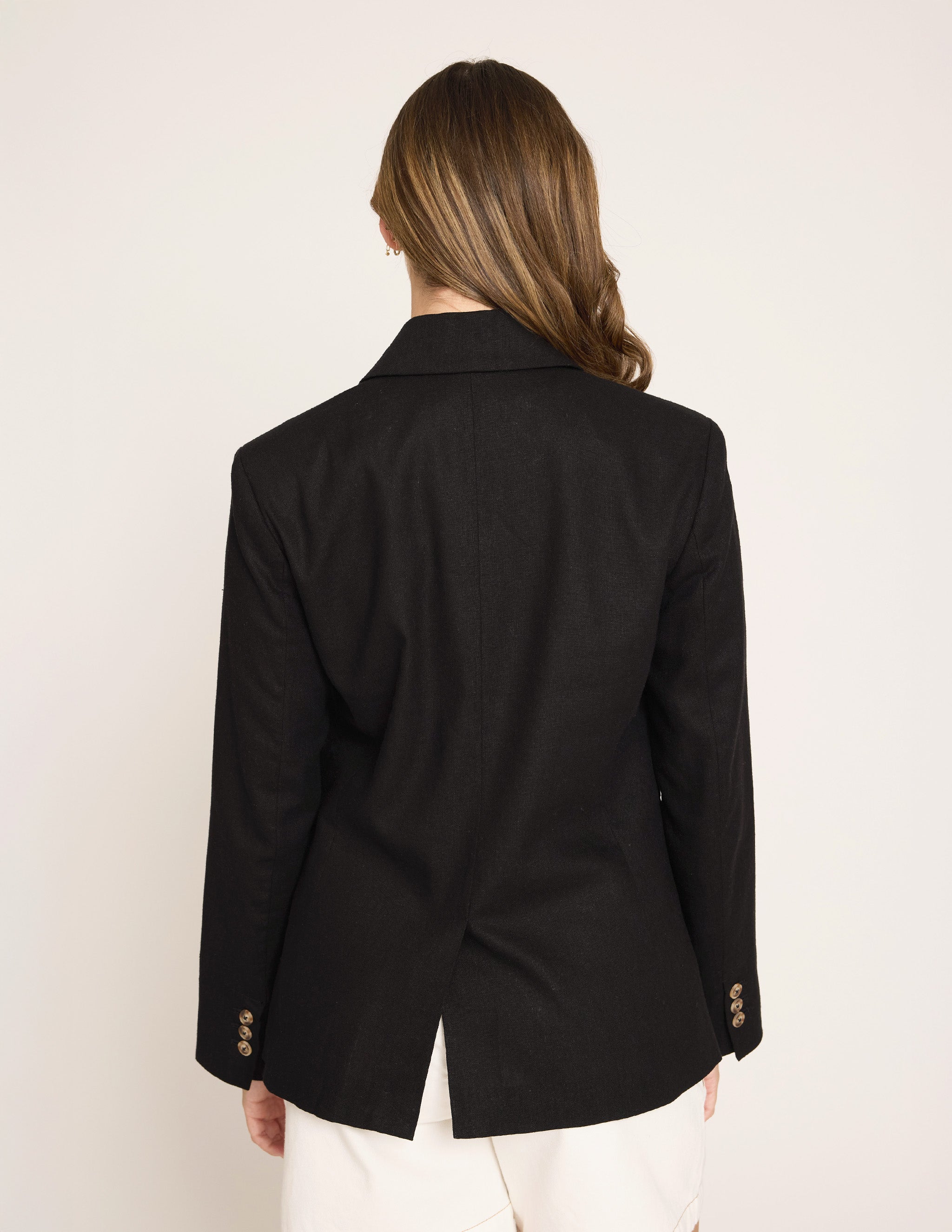 A woman viewed from the back wearing a black tailored blazer with gold-tone buttons on the cuffs and white pants, against a plain light background.