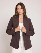 A woman wearing a dark brown pinstriped blazer over a white top and white pants, standing against a light gray studio background.