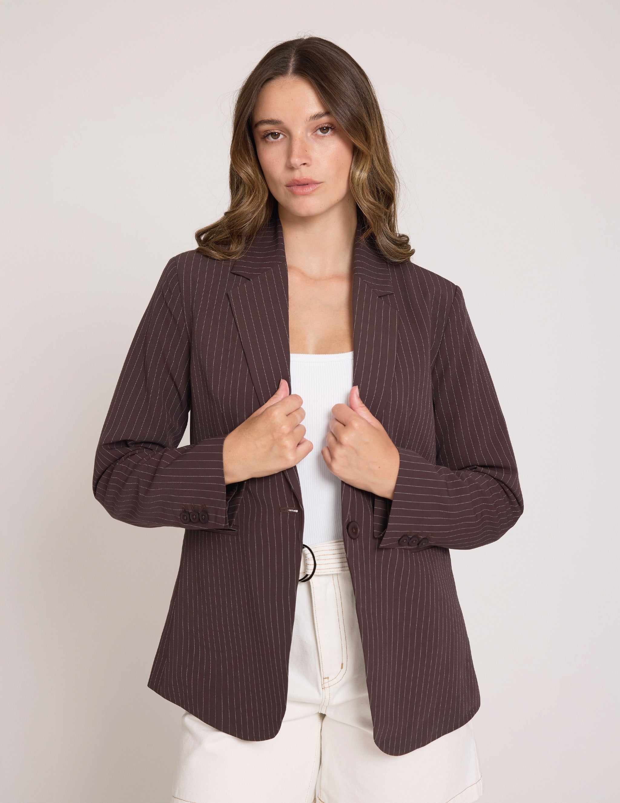 A woman wearing a dark brown pinstriped blazer over a white top and white pants, standing against a light gray studio background.