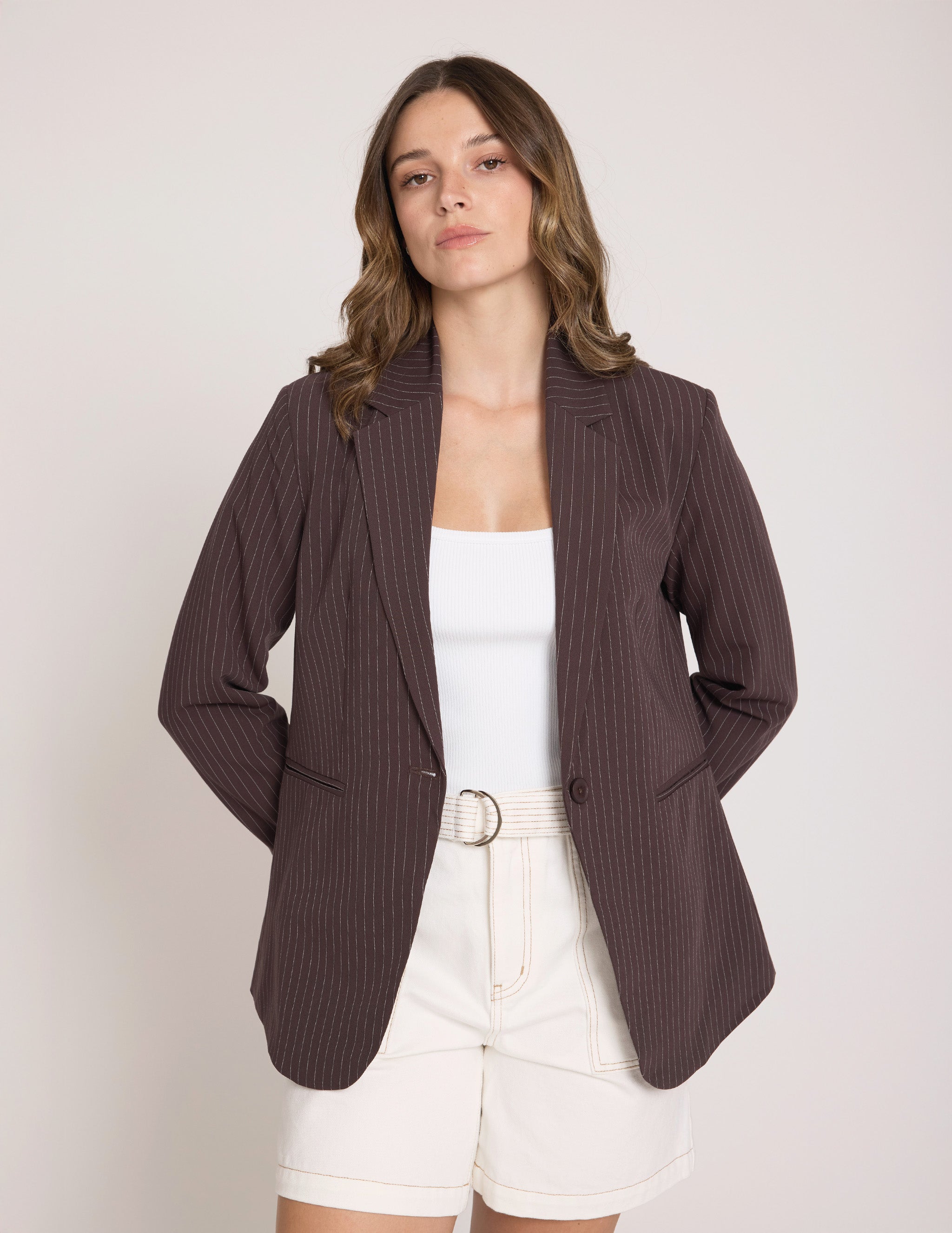 A woman wearing a dark brown ribbed pinstripe blazer over a white tank top and white shorts, standing against a light beige studio background.