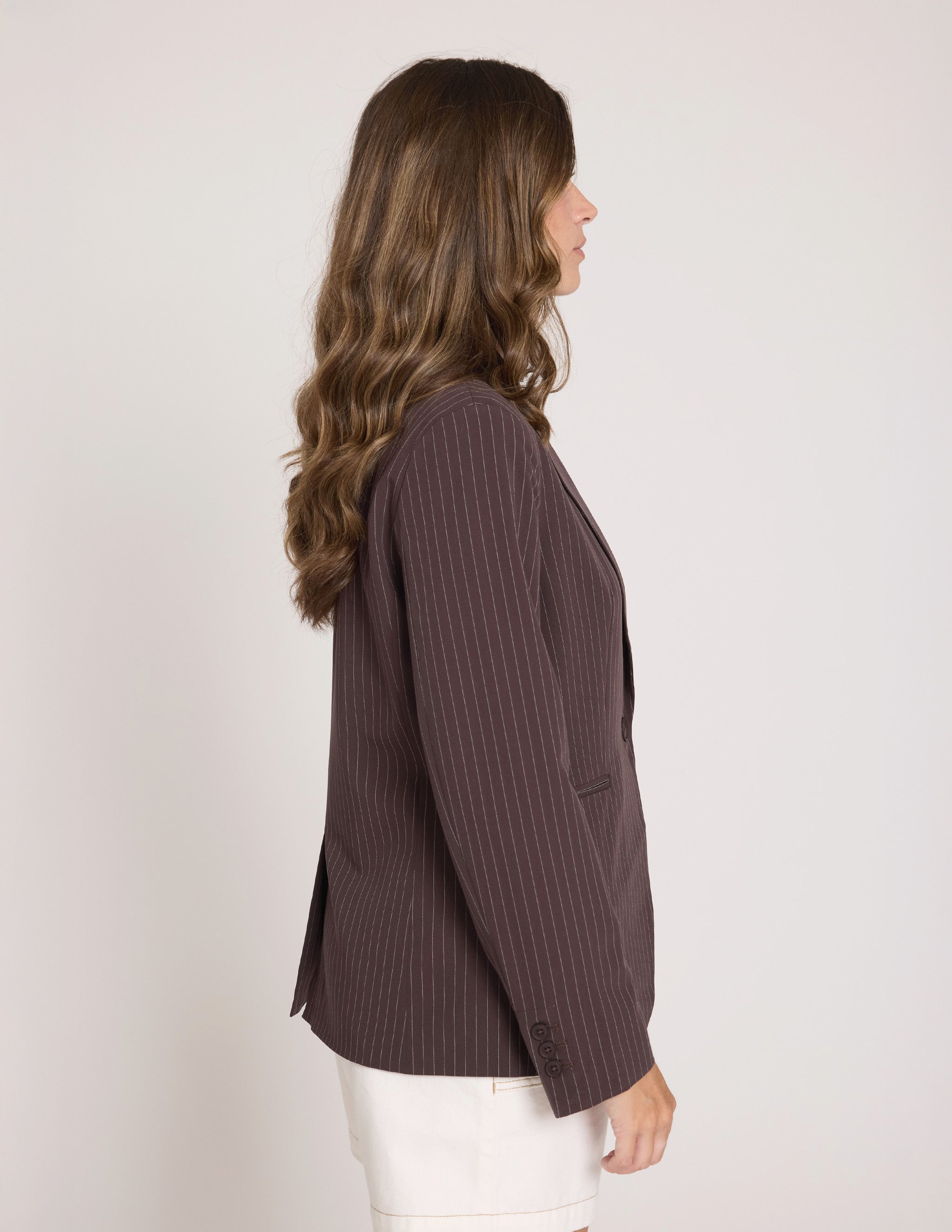A woman shown in side profile wearing a dark brown pinstriped blazer and white pants against a plain light background.
