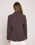 A woman viewed from the back wearing a dark brown pinstriped blazer and white pants against a plain light gray studio background.