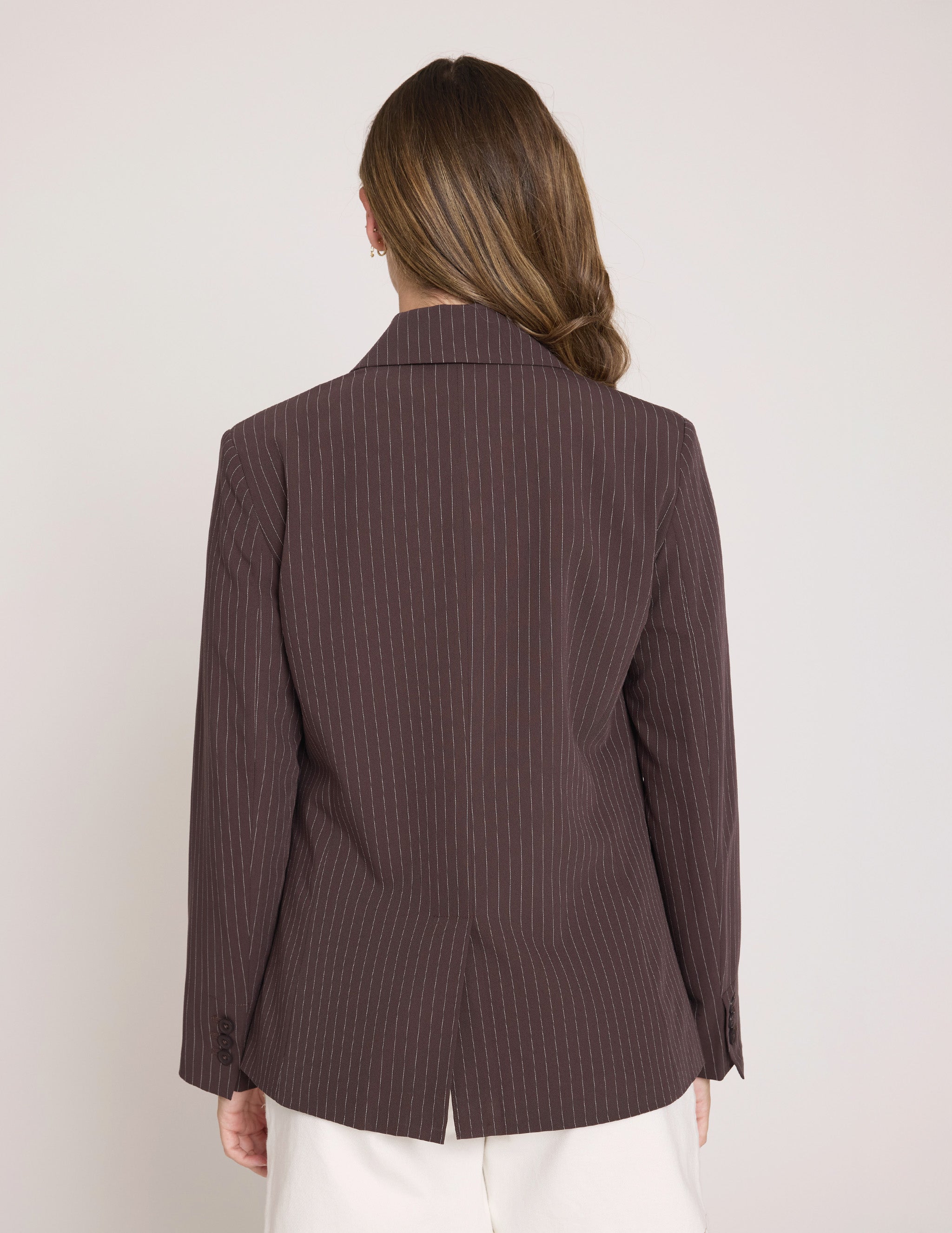 A woman viewed from the back wearing a dark brown pinstriped blazer and white pants against a plain light gray studio background.