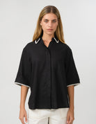 A woman modeling a black short-sleeve button-up shirt with white contrast trim on the collar and sleeve cuffs, paired with white pants, against a light gray studio background.