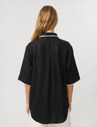 A woman viewed from the back wearing a black short-sleeve shirt with white scalloped trim on the collar and sleeve cuffs, paired with white bottoms, against a light gray studio background.