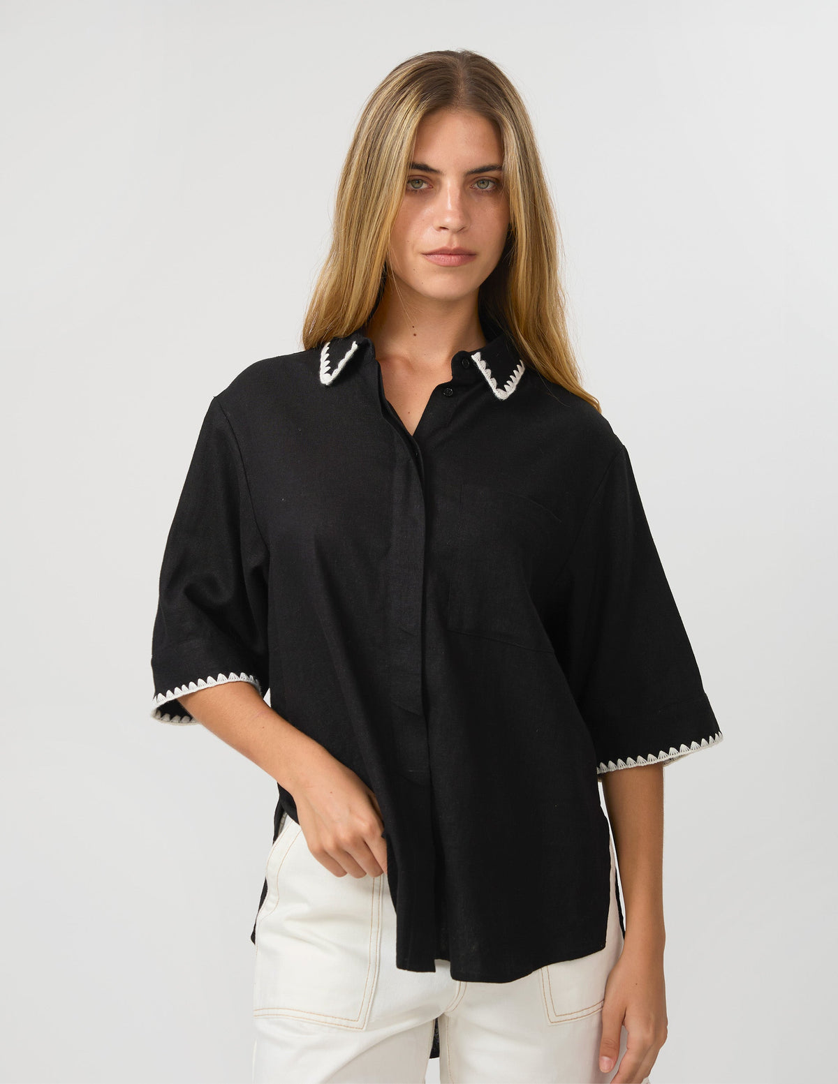 A woman modeling a black short-sleeve button-up shirt with white embroidered trim on the collar and sleeve cuffs, paired with white pants, against a light gray studio background.