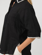 A close-up of a person wearing a black short-sleeve button-up shirt with white contrast stitching along the collar and sleeve cuffs, paired with white pants against a light gray background.