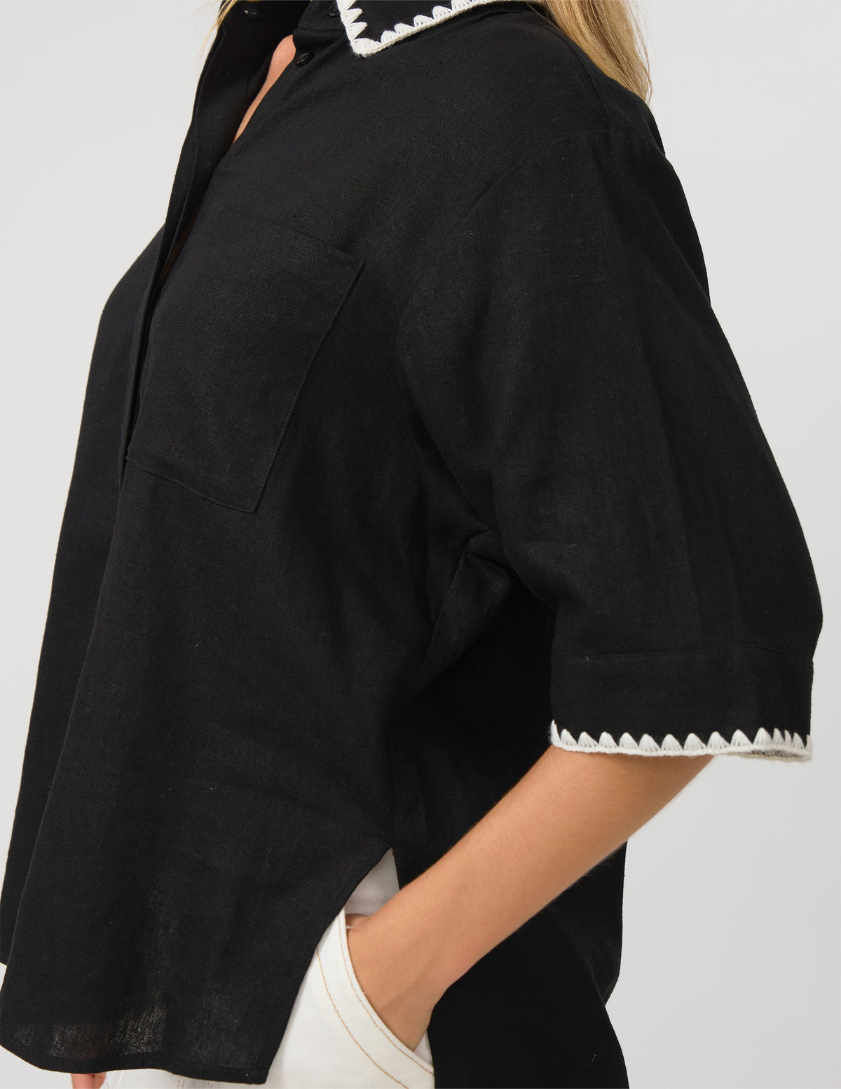 A close-up of a person wearing a black short-sleeve button-up shirt with white contrast stitching along the collar and sleeve cuffs, paired with white pants against a light gray background.