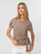 A woman modeling a short-sleeve leopard print top with a side tie, paired with white pants, against a plain light gray background.