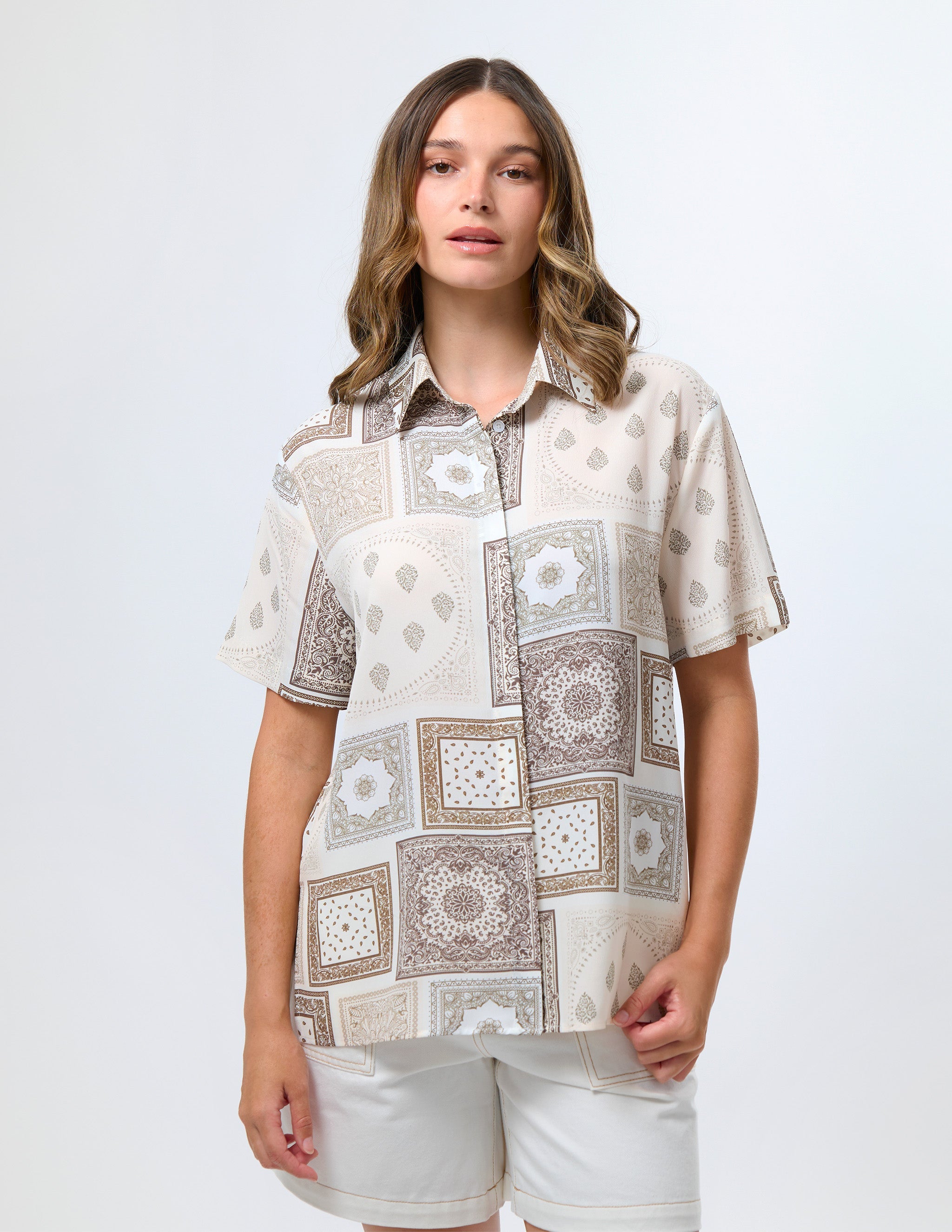 A woman modeling a short-sleeve button-up shirt with a beige, brown, and white geometric patchwork pattern, paired with white shorts, against a light gray studio background.